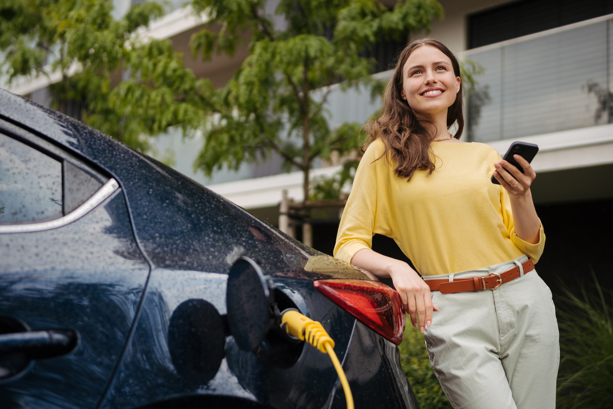 Junge Frau mit Smartphone wartet, während ihr Elektroauto an der Ladestation zu Hause aufgeladen wird – ein nachhaltiges und wirtschaftliches Verkehrskonzept. Junge Frau mit Smartphone wartet, während ihr Elektroauto geladen wird.